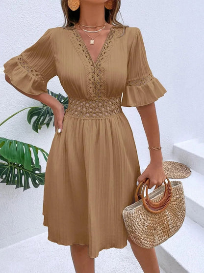 Woman wearing a camel brown V-neck mini dress with textured vertical striping, crochet lace detailing along the neckline, cinched lace waist, flounce sleeves, and accessorized with gold layered necklaces, statement earrings, and a woven handbag.