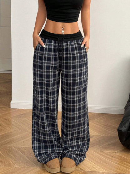 Front view of a woman wearing high-waisted navy blue and white plaid straight-leg, wide-leg sweatpants featuring a black elastic contrast waistband, styled with a black crop top and tan slip-on shoes.