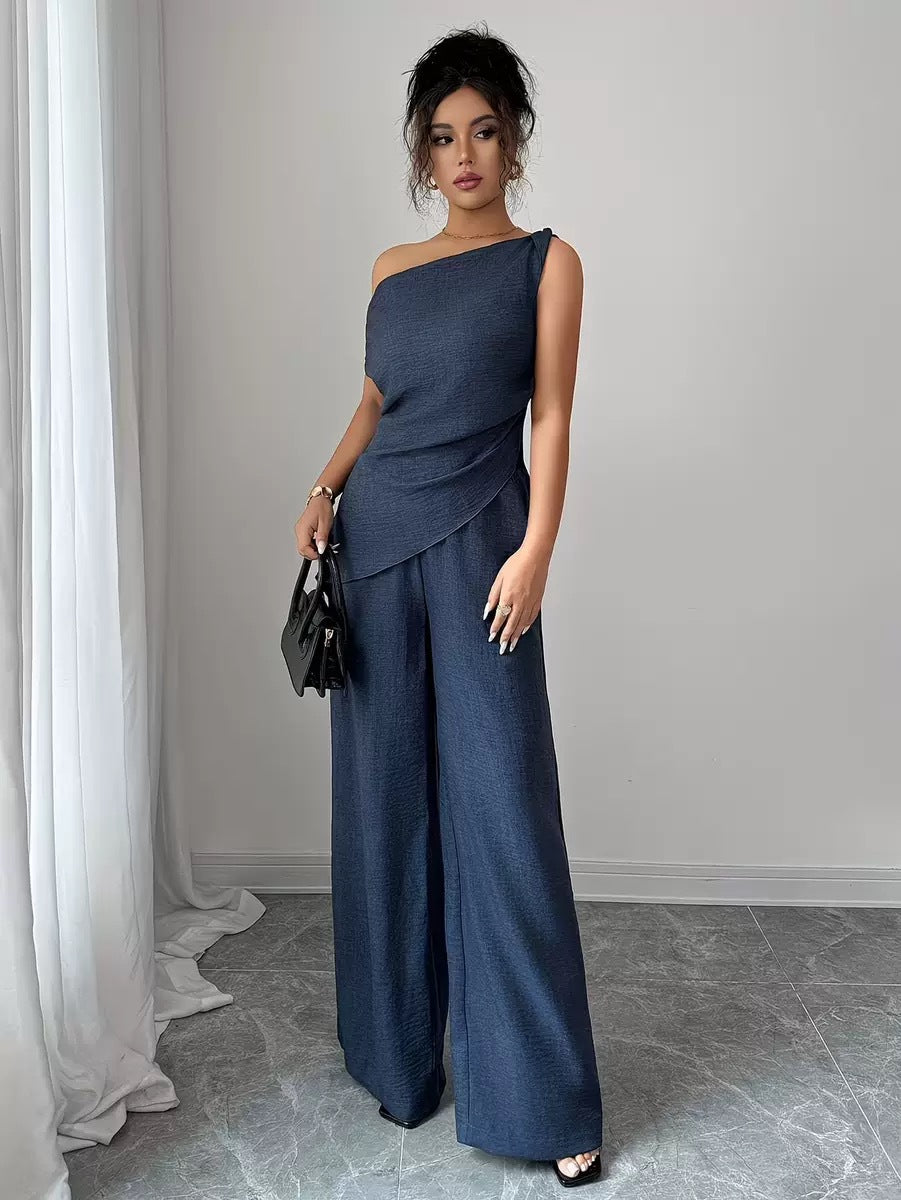 Woman wearing a navy blue casual two-piece set featuring a draped off-the-shoulder top with a diagonal front fold and matching high-waisted wide-leg pants, holding a black handbag.