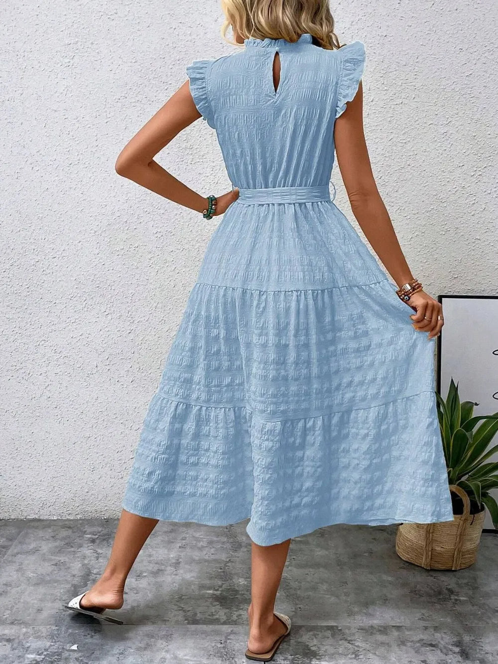 Back view of model wearing a light blue ruffled cap sleeve midi dress with a tied waist and white slide sandals