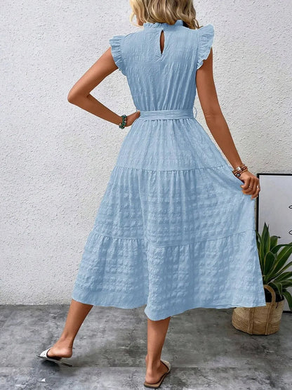 Back view of model wearing a light blue ruffled cap sleeve midi dress with a tied waist and white slide sandals