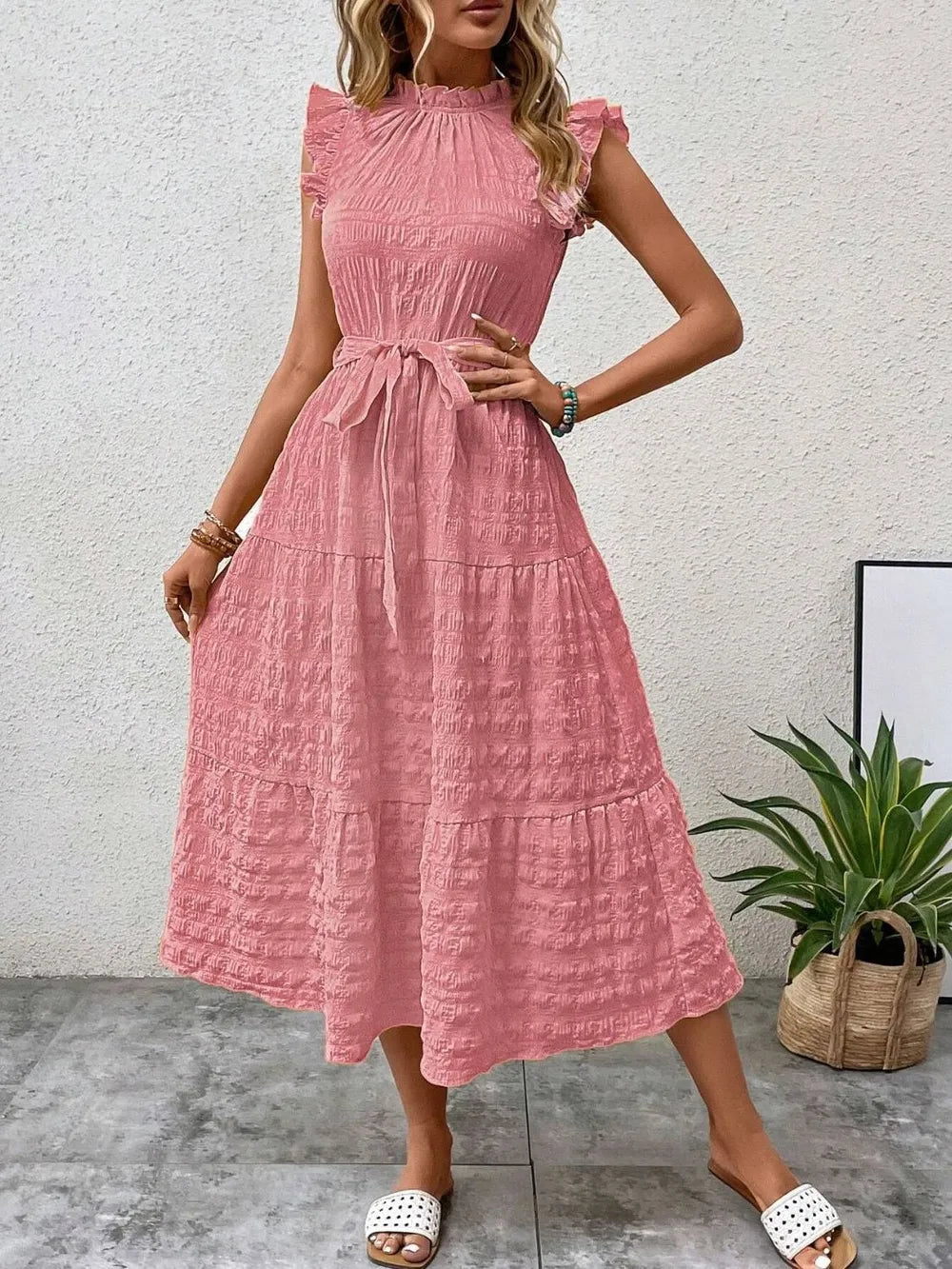 Front view of a model wearing a pink burnt coral ruffled cap sleeve midi dress with a tied waist, high neckline, and white slide sandals.