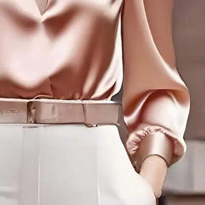 Close-up of a woman wearing a light pink satin V-neck long-sleeve blouse tucked into white trousers accented with a matching pink belt.