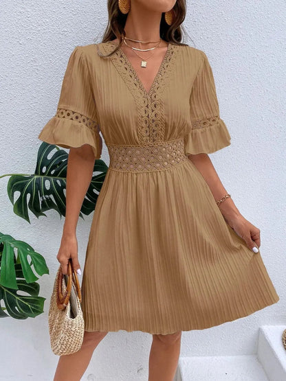 Woman wearing a camel brown V-neck mini dress with textured vertical striping, featuring crochet lace trim along the V-neckline, waist cutouts, and flounce sleeve cuffs, holding a woven straw tote bag.
