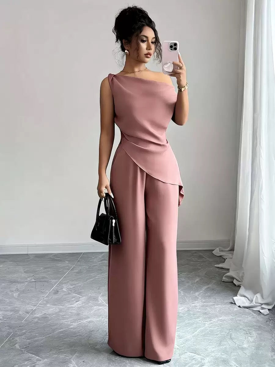 Woman taking a mirror selfie wearing a dusty pink casual two-piece set featuring a draped off-the-shoulder top with a diagonal front fold and matching high-waisted wide-leg pants, holding a black handbag and smartphone.