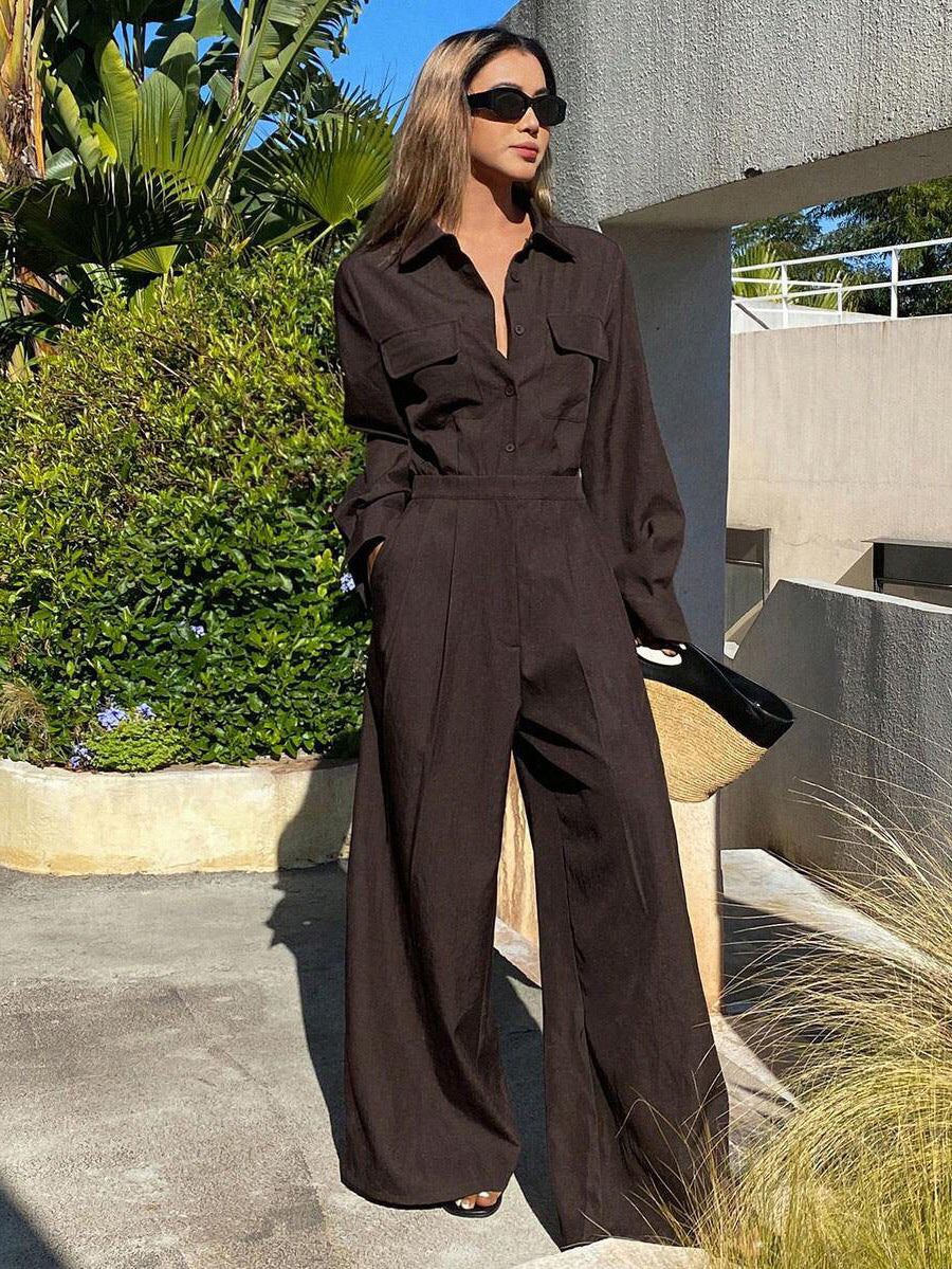 Full-length outdoor shot of a woman wearing a dark brown collared long-sleeve jumpsuit with chest flap pockets, sunglasses, and wide-leg trousers while carrying a straw handbag.