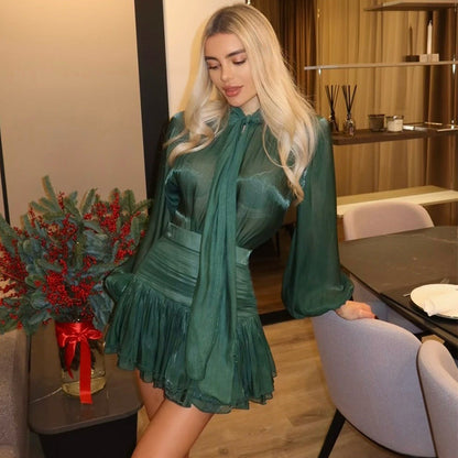 Woman wearing an emerald green high-waisted two-piece set featuring a long-sleeve blouse with a tied neck tie accent and a ruched, layered mini skirt, posing in an interior setting near a dining table