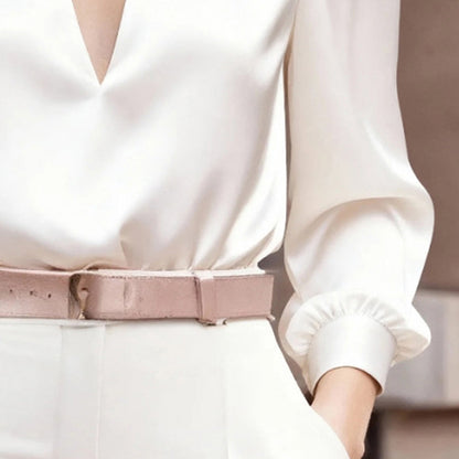 Close-up of a woman wearing a white satin V-neck long-sleeve blouse tucked into white trousers accented with a matching beige belt.