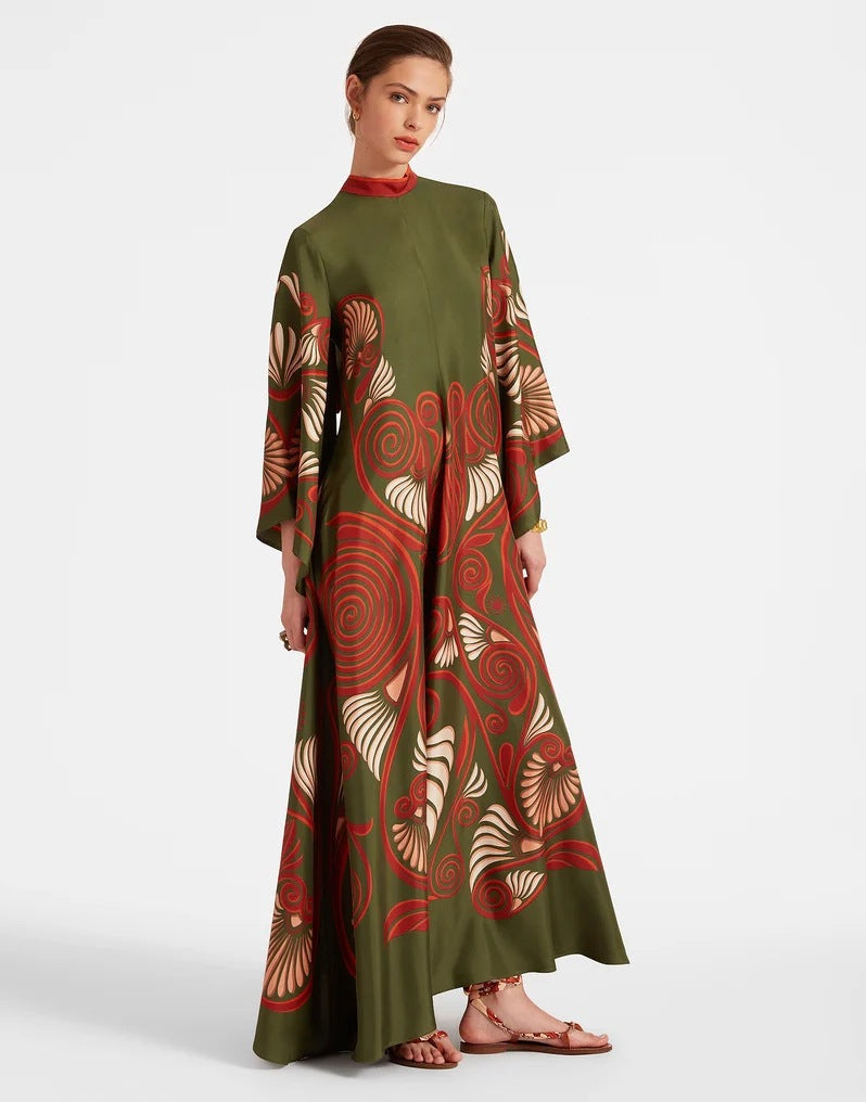 Full-length model shot of an olive green maxi dress featuring an abstract rust-red and cream floral pattern, high red stand collar, wide flared 3/4 sleeves, and a sweeping A-line silhouette.