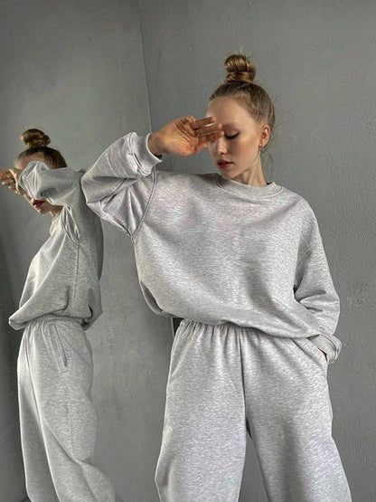 Woman wearing a gray loose-fit crewneck sweatshirt and matching jogger pants two-piece set, posed with her hand near her head, showing a side-by-side or reflective view.