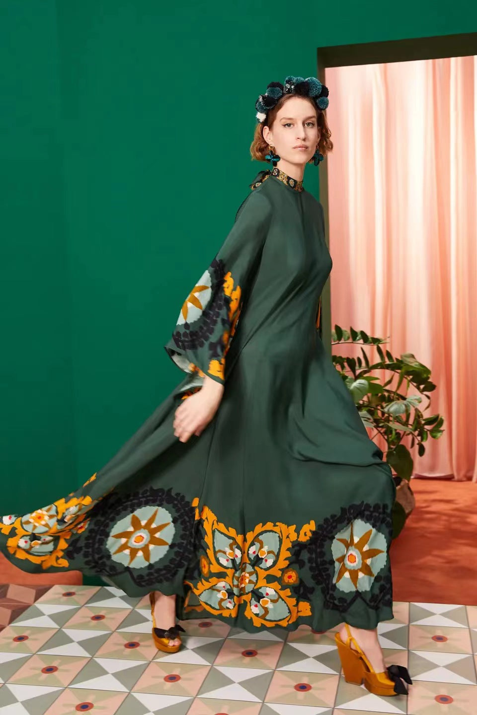 Full-length model shot of a dark green maxi dress featuring wide statement sleeves with mustard and blue tile print accents, a wide ornate hem border, high neckline, and sweeping A-line silhouette.