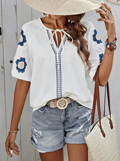 Woman wearing a white V-neck blouse featuring blue embroidered floral sleeves, a braided tie-neck, and patterned vertical trim, styled with distressed cuffed denim shorts, a woven round-buckle belt, a straw hat, and holding a tote bag.