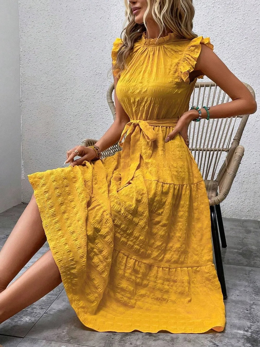 Model sitting on a woven chair wearing a gold yellow ruffled cap sleeve midi dress with a tied waist