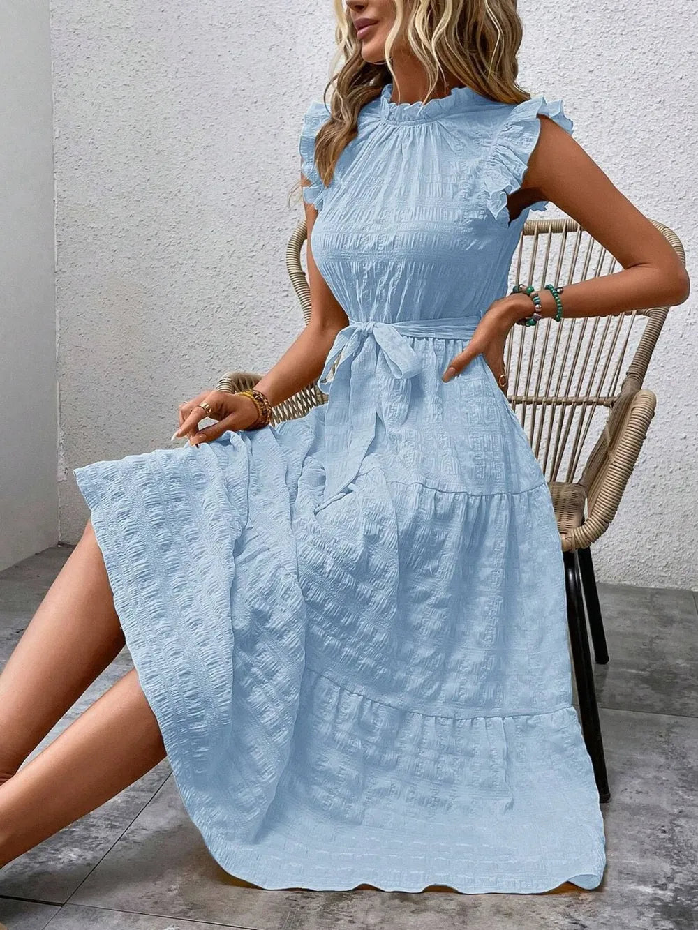 Model sitting on a woven chair wearing a light blue ruffled cap sleeve midi dress with a tied waist