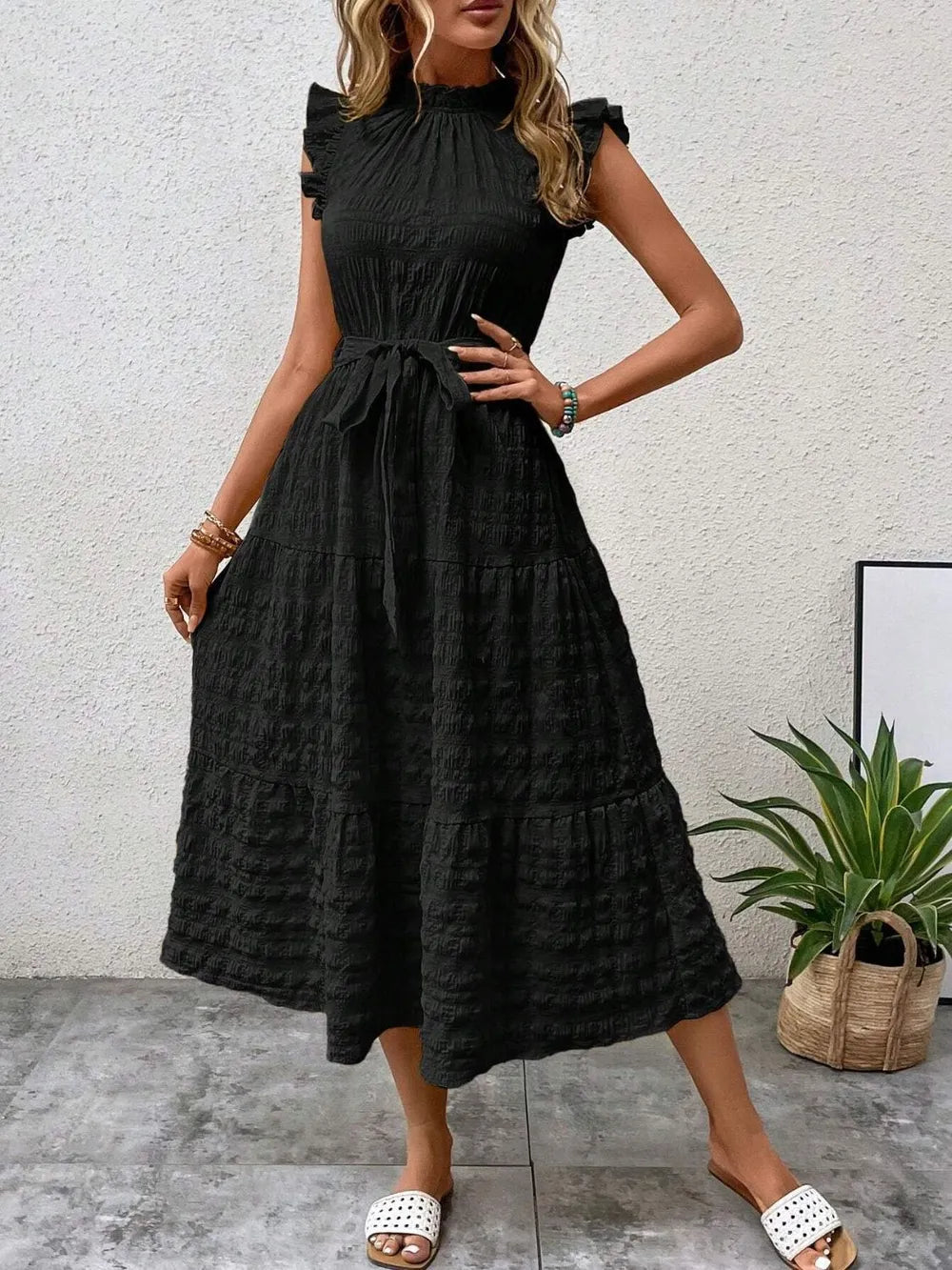 Front view of model wearing a black ruffled cap sleeve midi dress with a tied waist and white slide sandals