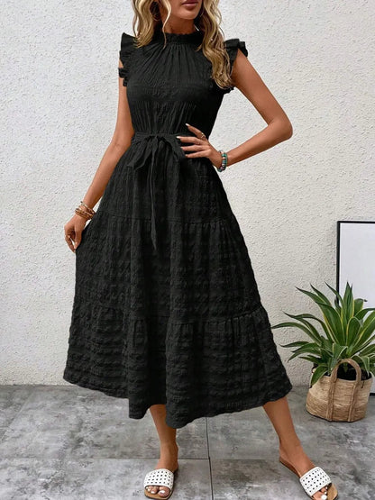 Front view of model wearing a black ruffled cap sleeve midi dress with a tied waist and white slide sandals