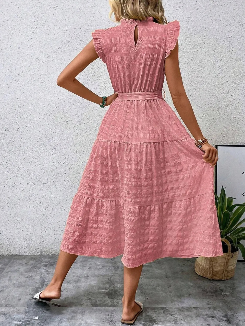 Back view of a model wearing a pink burnt coral ruffled cap sleeve midi dress with a tied waist, high neckline with keyhole detail, and white slide sandals.
