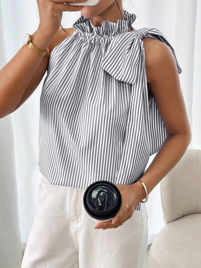 Front-facing view of a model holding a black cup while wearing a purple and white striped sleeveless top with a ruffled high neck and side tie-knot bow.