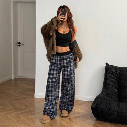 Full-length view of a woman wearing high-waisted navy blue, white, and grey plaid wide-leg lounge pants with a black elastic contrast waistband, styled with a black cropped tank top, a brown faux fur jacket, layered necklaces, and tan platform boots.