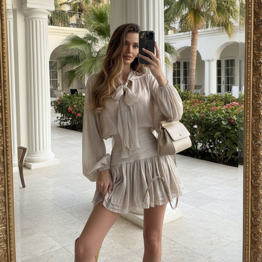 Mirror selfie of a woman wearing a light khaki high-waisted two-piece set featuring a long-sleeve blouse with a tied neck tie accent and a ruched, layered mini skirt, styled with a shoulder bag.