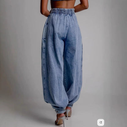 Back view of high-waisted light blue denim harem pants featuring an elastic waistband, relaxed loose-fit legs, and cinched ankle cuffs paired with heeled sandals.