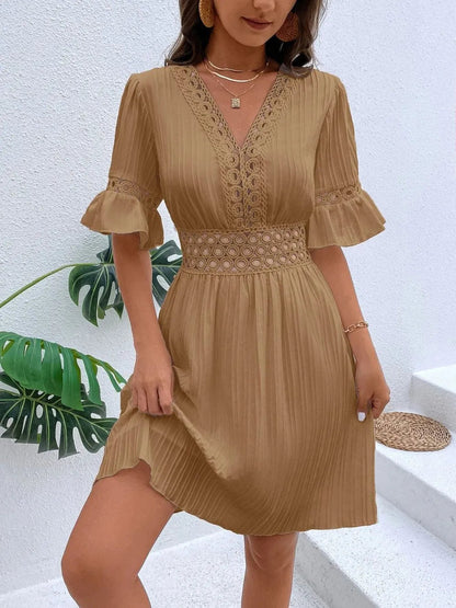 Woman wearing a camel tan V-neck mini dress with textured vertical striping, featuring crochet lace trim along the neckline, a cinched lace waistband, flounce sleeves, and layered gold necklaces.