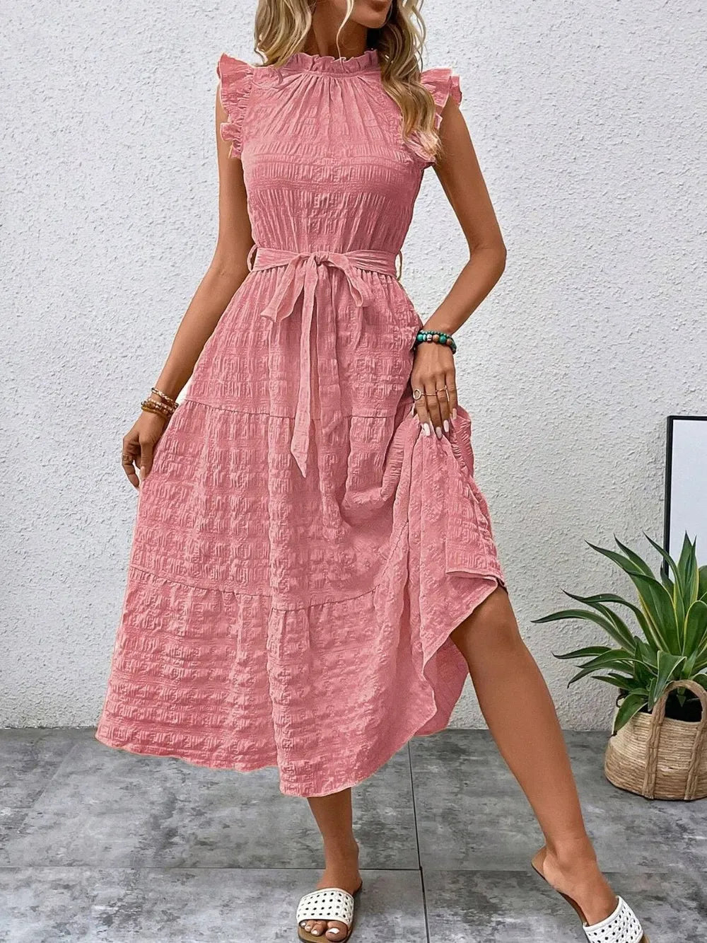 Front view of a model stepping forward wearing a pink burnt coral ruffled cap sleeve midi dress with a tied waist, high neckline, and white slide sandals.
