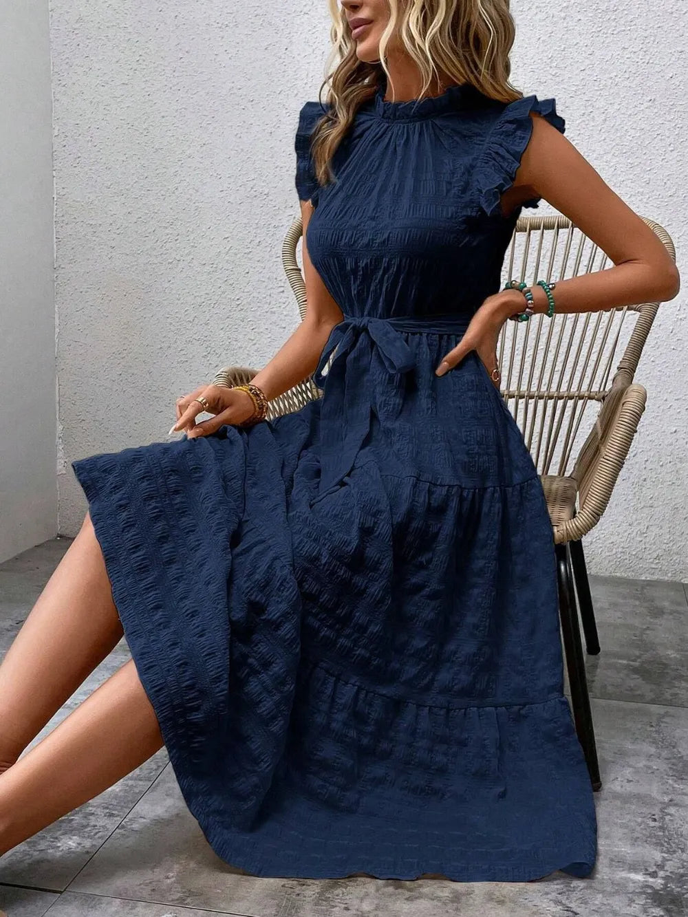 Model sitting on a woven chair wearing a dark blue ruffled cap sleeve midi dress with a tied waist.