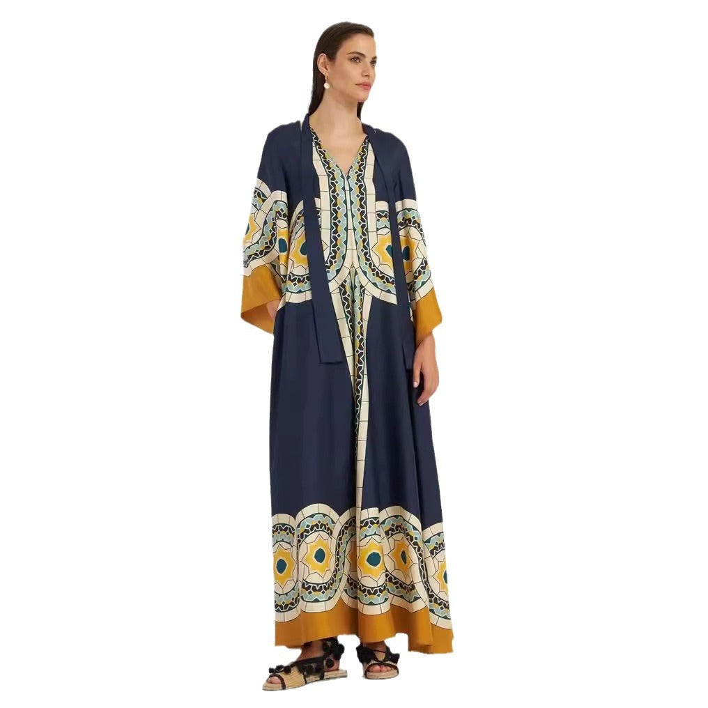 Full-length model shot of a dark navy blue maxi dress featuring a circular tile and botanical print in yellow and white, wide mustard-yellow sleeve cuffs, decorative vertical border trim, and a split V-neckline.