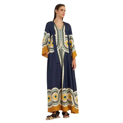 Full-length model shot of a dark navy blue maxi dress featuring a circular tile and botanical print in yellow and white, wide mustard-yellow sleeve cuffs, decorative vertical border trim, and a split V-neckline.