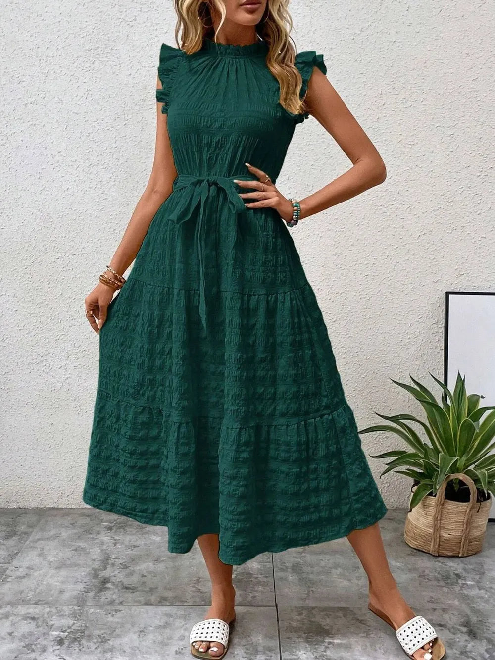 Front view of model wearing a dark green ruffled cap sleeve midi dress with a tied waist and white slide sandals