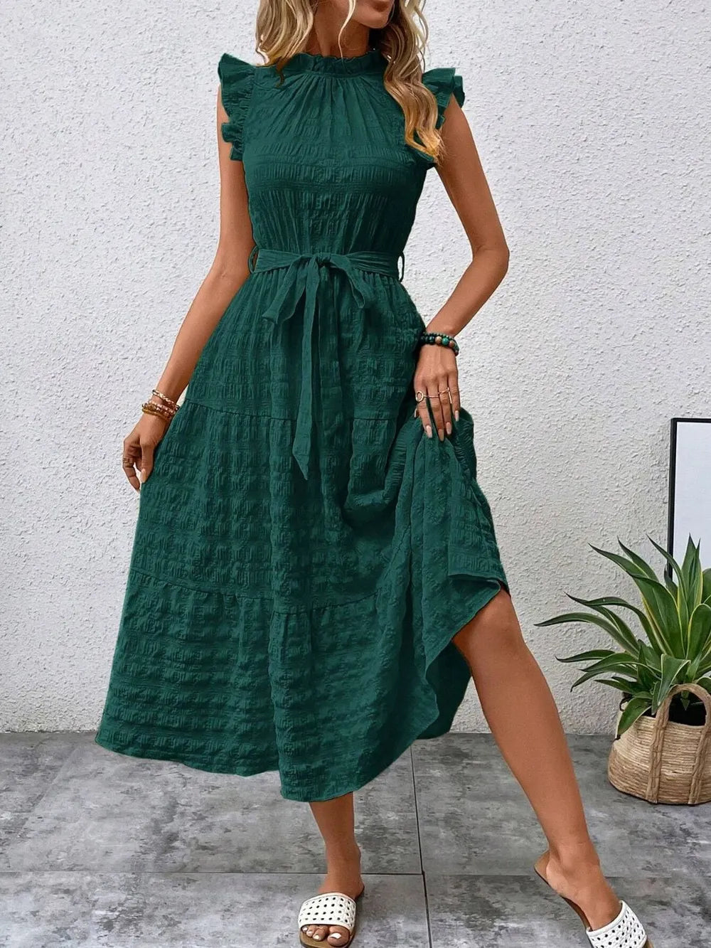 Front view of model stepping forward wearing a dark green ruffled cap sleeve midi dress with a tied waist and white slide sandals