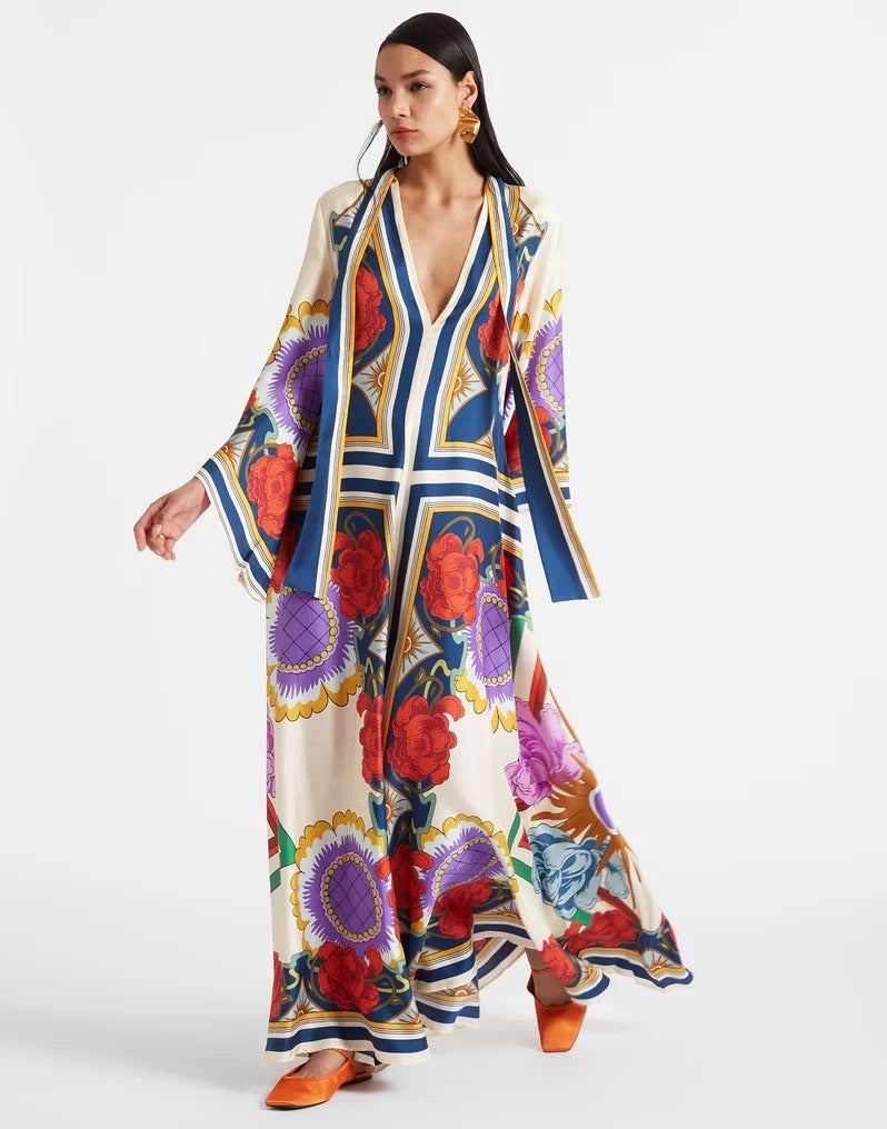 Women's floor-length A-line maxi dress in off-white featuring a vibrant botanical and floral pattern with blue border accents, a plunging V-neckline, and wide 3/4 flared sleeves.