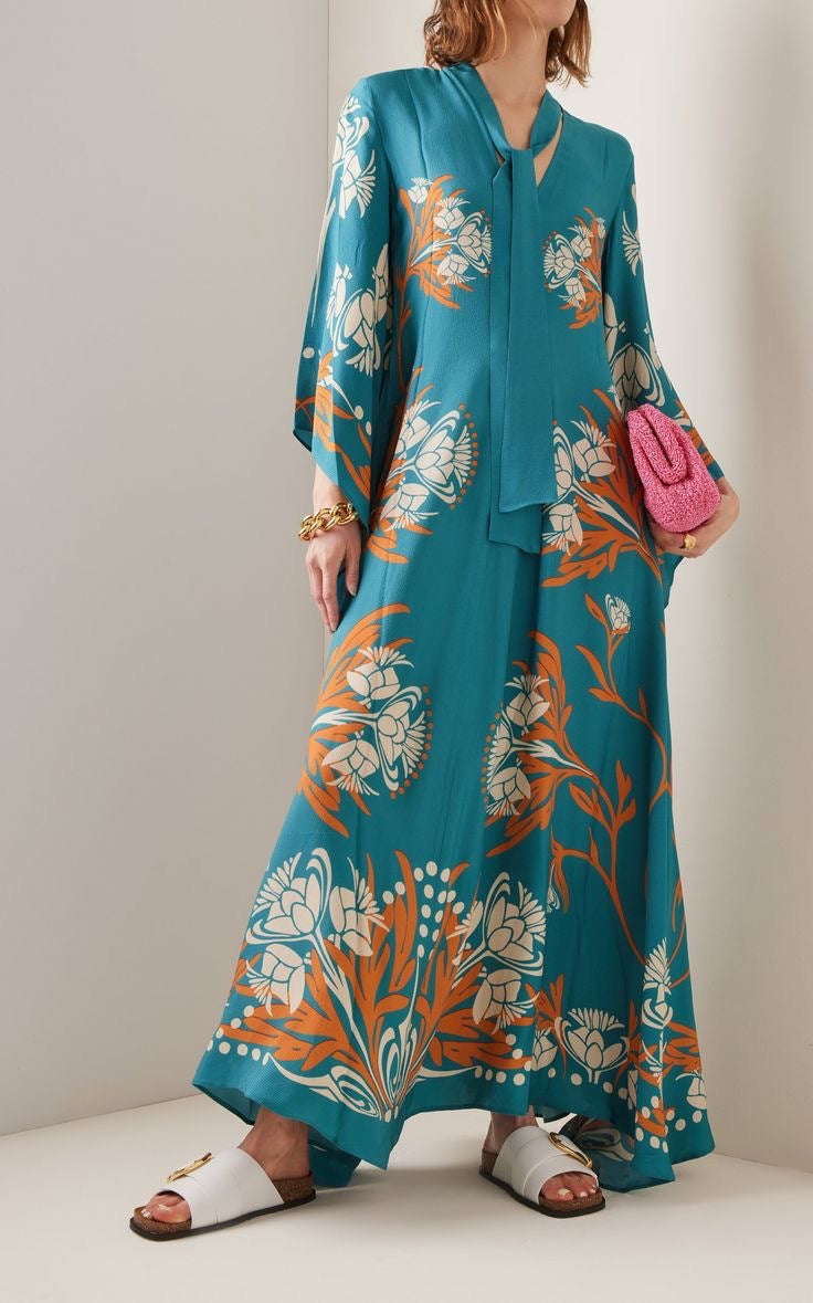 Full-length model shot of a teal blue maxi dress featuring an orange and white floral branch print, a high neckline with a matching neck tie scarf, and wide long flared sleeves