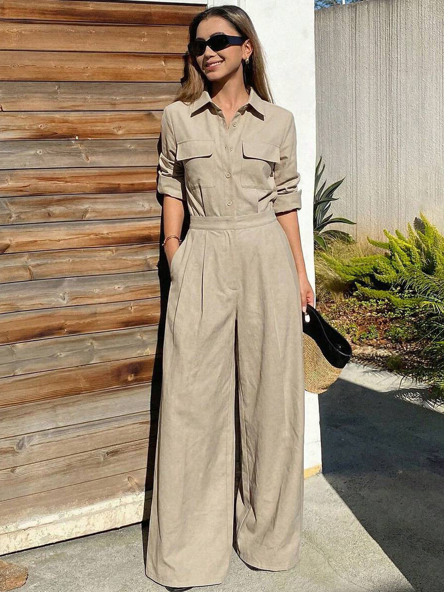 Full-length outdoor shot of a woman wearing an olive green collared long-sleeve jumpsuit with chest flap pockets, sunglasses, and wide-leg trousers while holding a straw clutch.