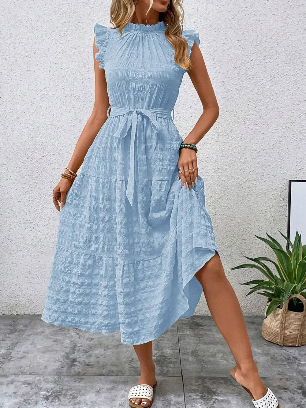 Front view of model stepping forward wearing a light blue ruffled cap sleeve midi dress with a tied waist and white slide sandals