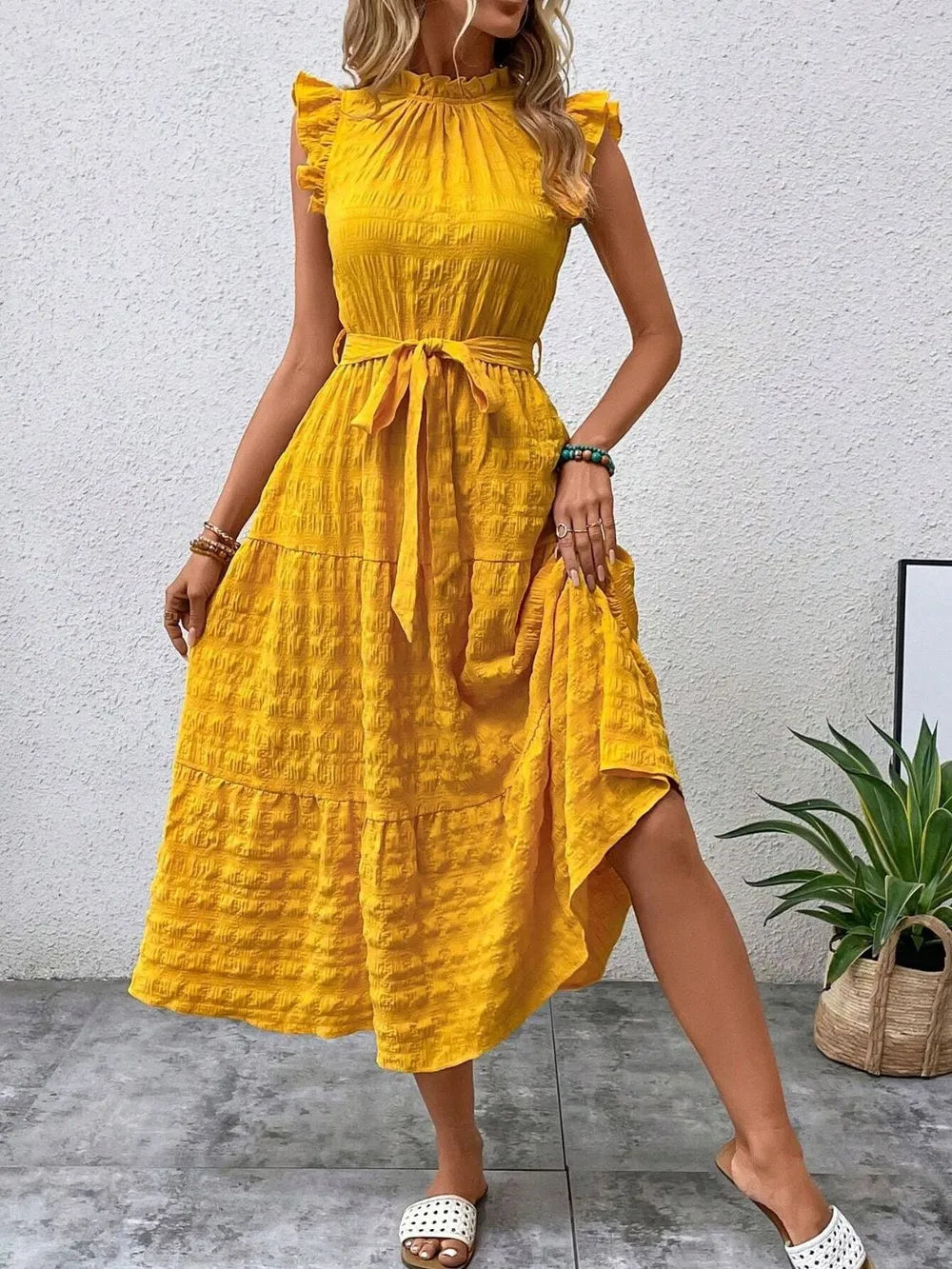 Front view of model stepping forward wearing a gold yellow ruffled cap sleeve midi dress with a tied waist and white slide sandals
