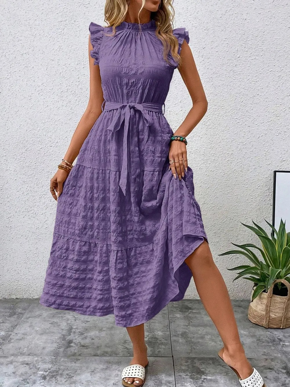 Front view of a model wearing a purple ruffled cap sleeve midi dress with a tied waist, high neckline, and white slide sandals.