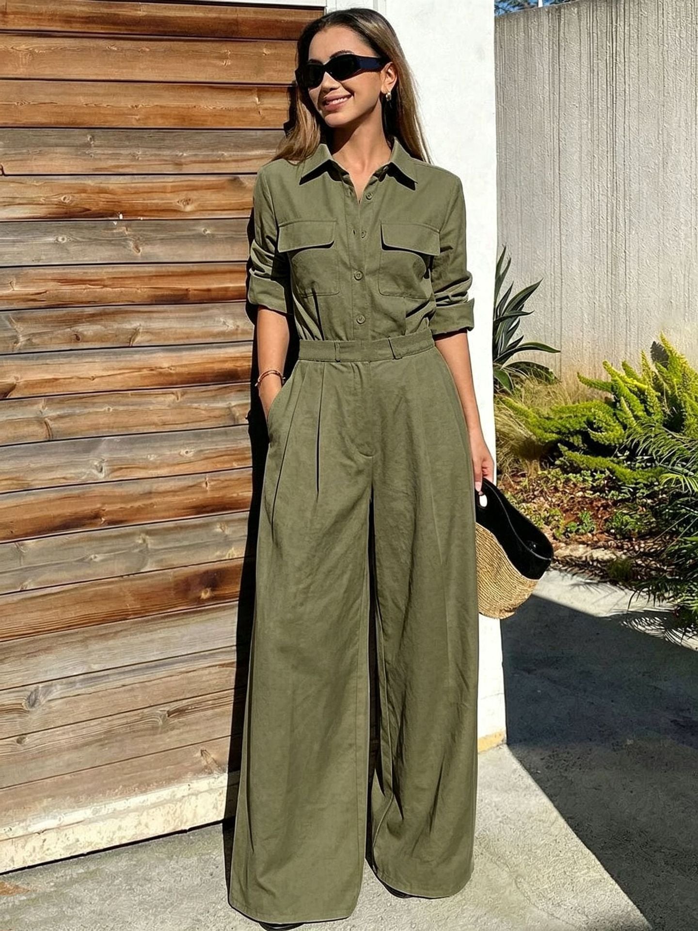 Full-length outdoor shot of a woman wearing an olive green collared long-sleeve jumpsuit with chest flap pockets, sunglasses, and wide-leg trousers while holding a straw clutch.