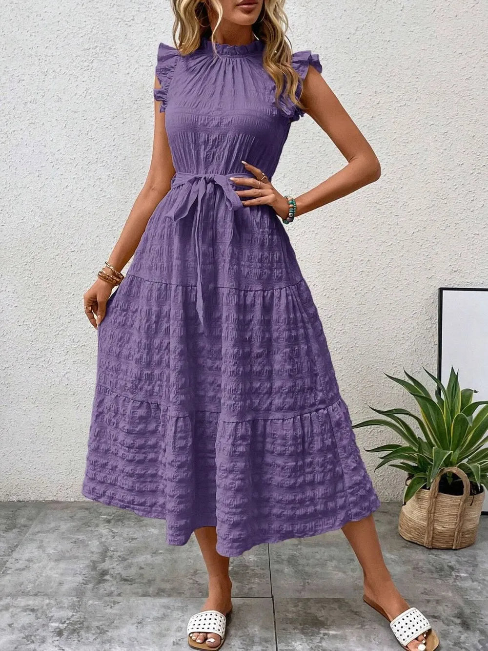 Front view of a model wearing a purple ruffled cap sleeve midi dress with a tied waist and white slide sandals.