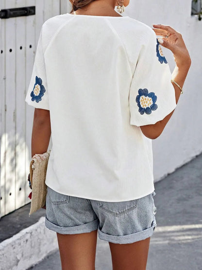 Back view of a woman wearing a loose white V-neck top with blue floral embroidered short sleeves and cuffed denim shorts