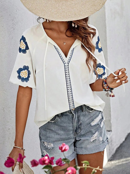 Woman wearing a cream V-neck blouse with blue embroidered flowers on short wide sleeves, geometric embroidered trim along the V-neckline and tie collar, paired with distressed light-wash denim shorts and a straw hat.