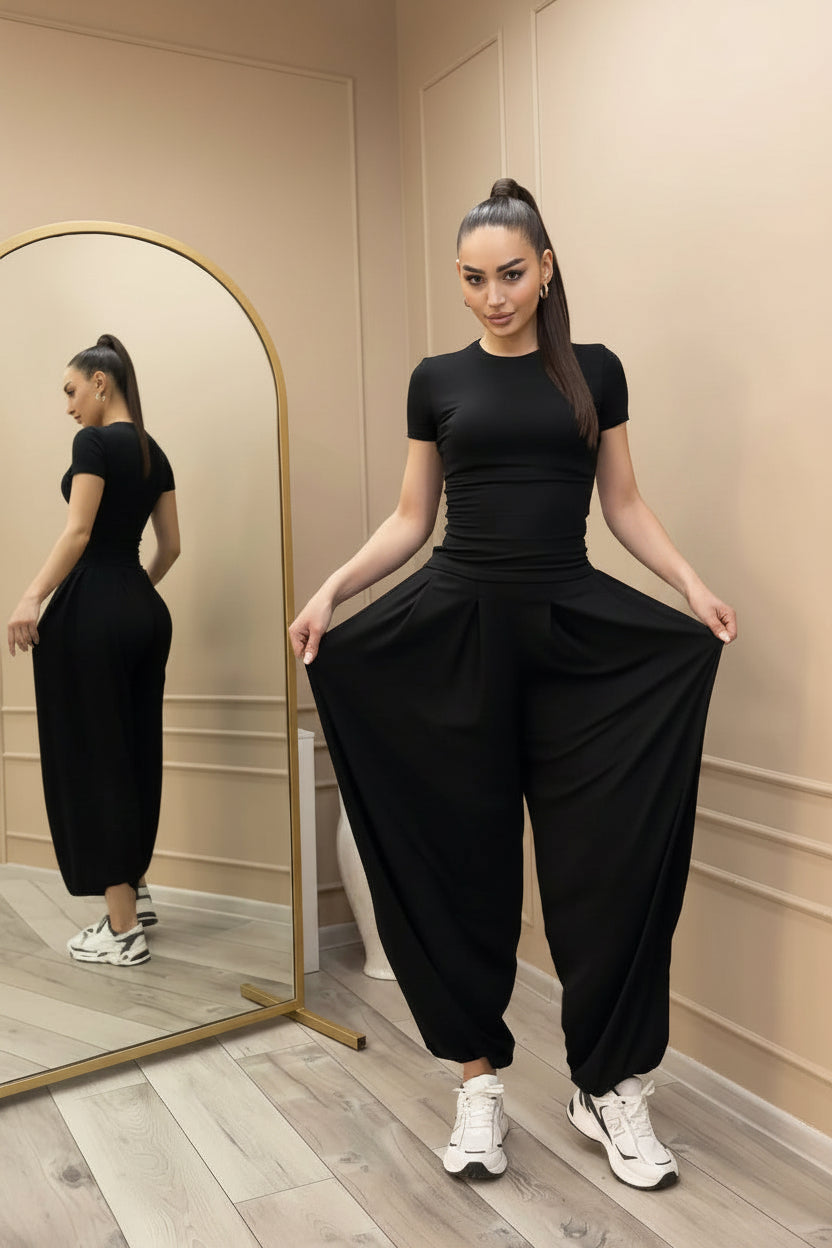 Model with a high ponytail posing in front of a mirror wearing a black short-sleeve fitted tee and matching volume harem pants paired with white sneakers.