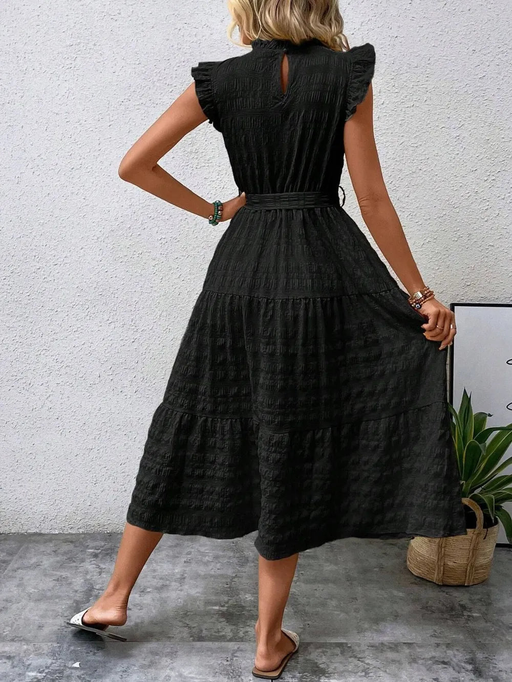 Back view of a model wearing a black ruffled cap sleeve midi dress with a tied waist, high neckline with keyhole detail, and white slide sandals.