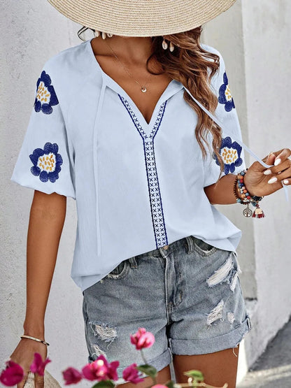 Front view of a woman wearing a straw hat and light blue V-neck top with blue floral embroidered sleeves, braided neck tie, and distressed denim shorts.