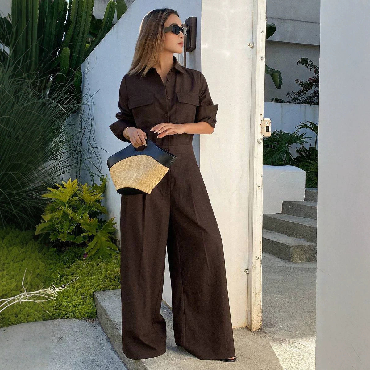 Full-length outdoor shot of a woman wearing a loose-fit, dark brown collared long-sleeve jumpsuit with chest flap pockets, sunglasses, and wide-leg trousers while holding a straw clutch.