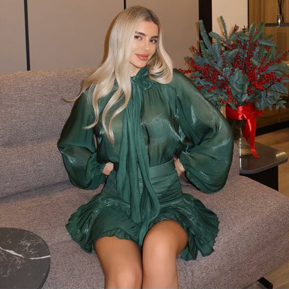 Blonde woman wearing an emerald green two-piece set featuring a sheer, long-sleeve blouse with a high-neck tie accent and a high-waisted ruched mini skirt, seated on a grey couch.