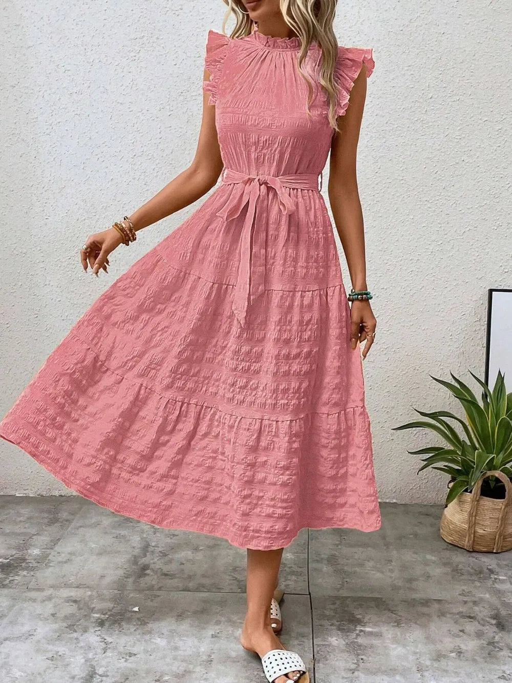 Front view of a model stepping forward wearing a pink burnt coral ruffled cap sleeve midi dress with a tied waist and white slide sandals.