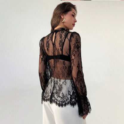 Back-angled view of a model wearing a sheer black lace hollow-out long sleeve top featuring a mock neck and white trousers.