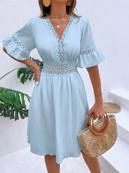 Woman wearing a light blue V-neck mini dress with textured vertical striping, featuring crochet lace trim along the V-neckline, waist cutouts, and flounce sleeves, styled with layered gold necklaces and holding a woven straw handbag.